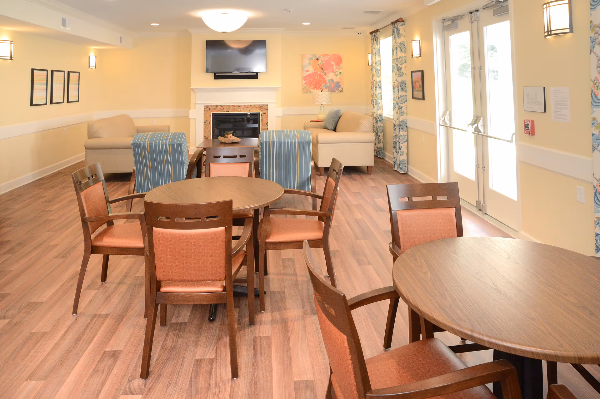 Bright communal room with round wooden tables and chairs, sofas near a fireplace and a wall-mounted TV.