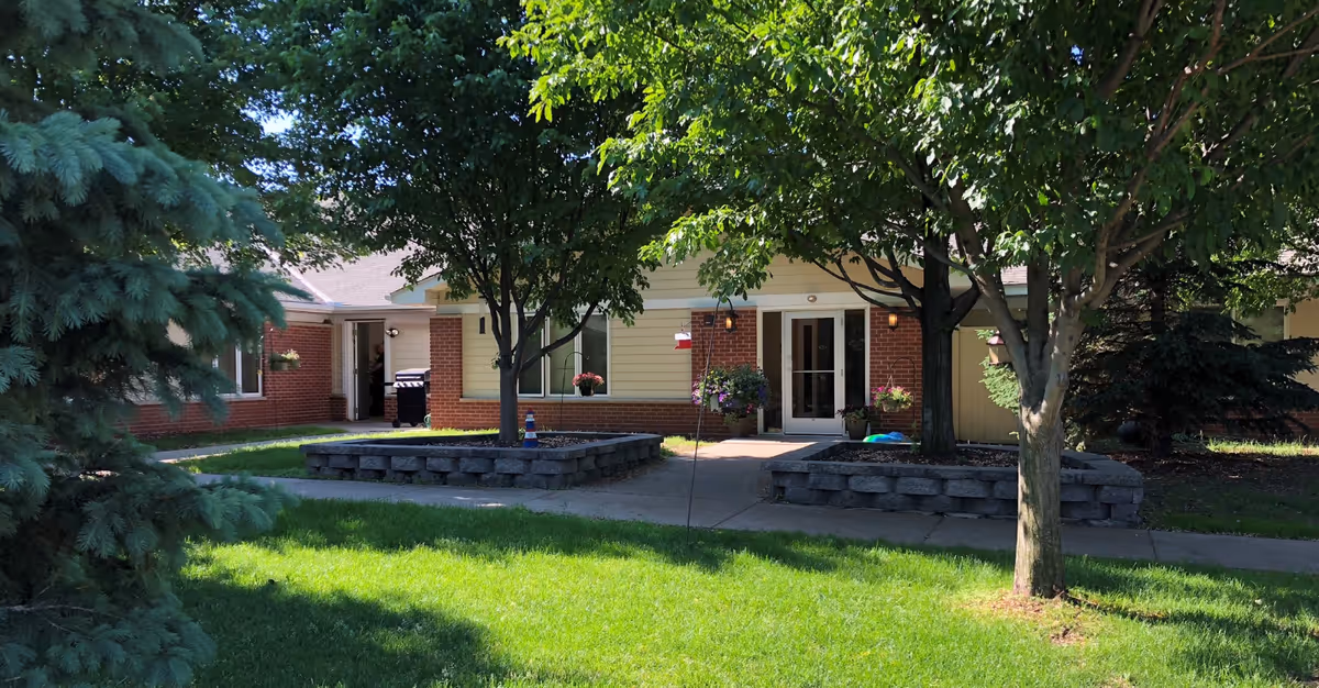 Exterior view of a senior living facility building with a brick and light yellow siding facade, surrounded by green grass, trees, and flower pots near the entrance. A person is visible near an open door on the left side.