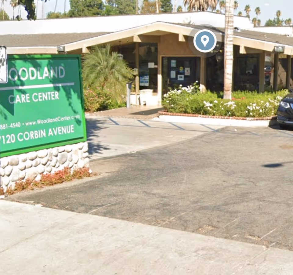Exterior view of Woodland Care Center showing the entrance with a covered walkway, surrounded by greenery and palm trees. A green sign with the facility name, website, phone number, and address is visible on the left side near the sidewalk.