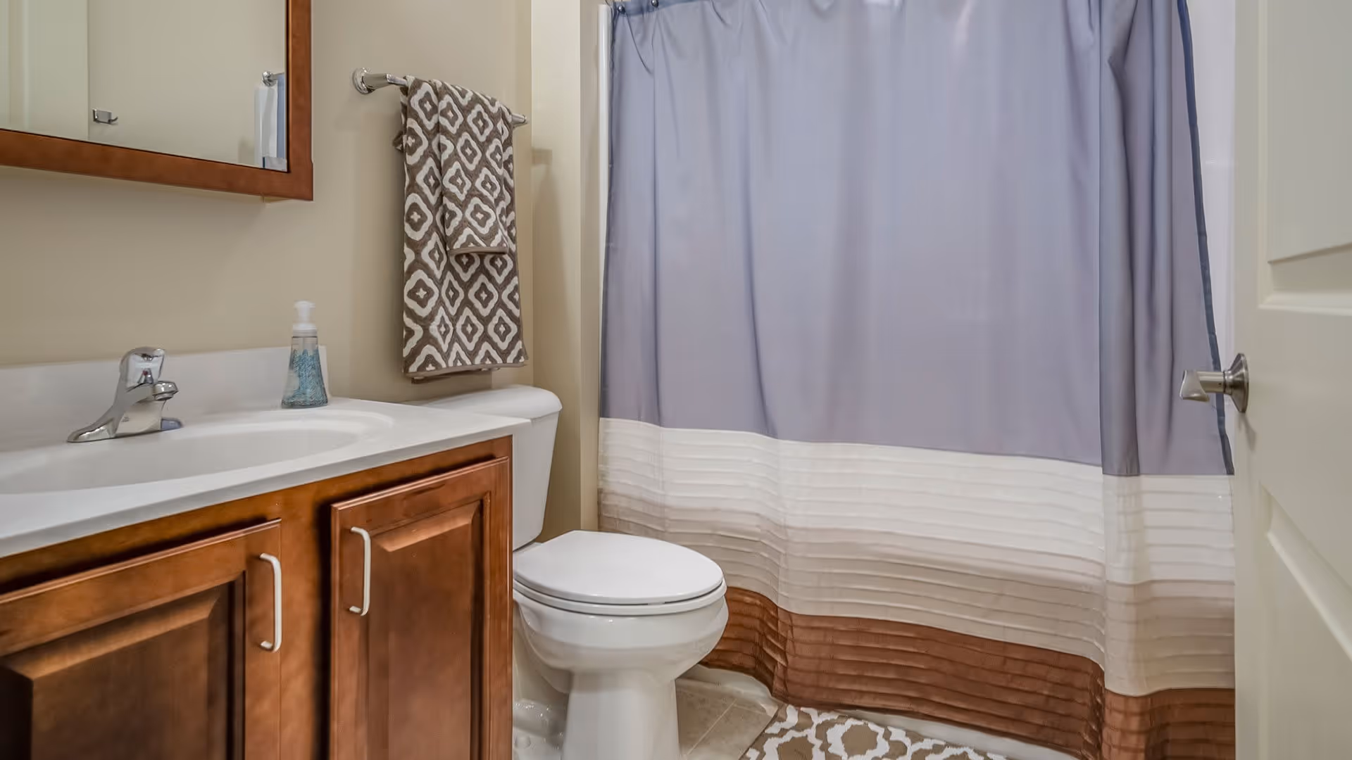 A bathroom with a wooden vanity cabinet, white sink, and chrome faucet. A patterned towel hangs on a towel rack above a white toilet. A shower with a curtain featuring gray, white, and brown horizontal stripes is visible. The bathroom door is partially open.