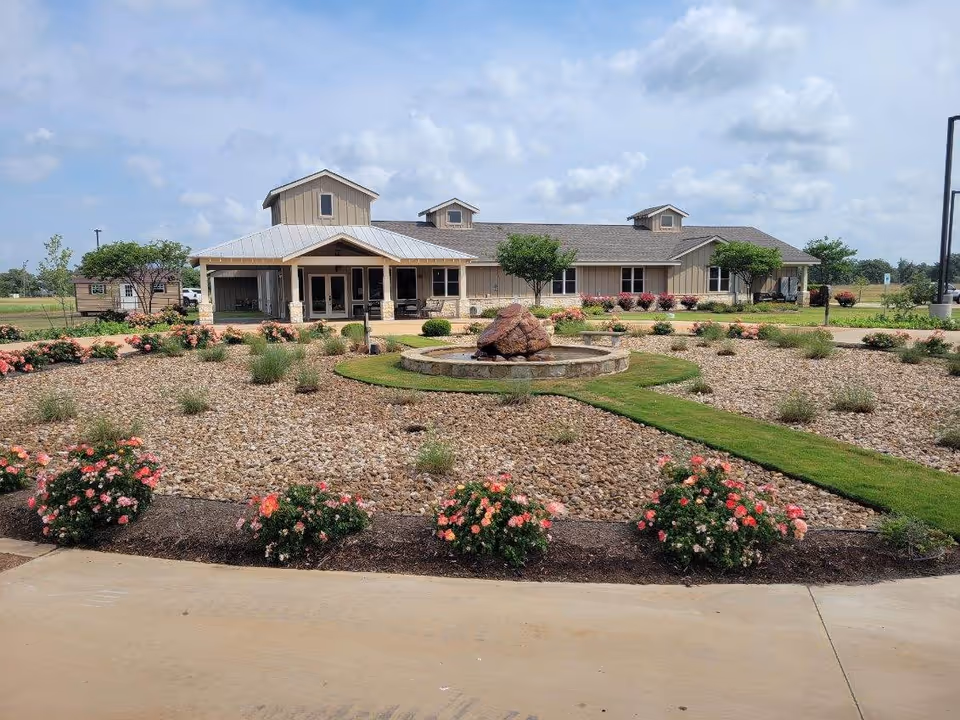 Exterior view of a single-story building with a metal roof and beige siding, surrounded by a landscaped garden featuring a circular stone water fountain, green grass paths, bushes, and flowering plants under a partly cloudy sky.