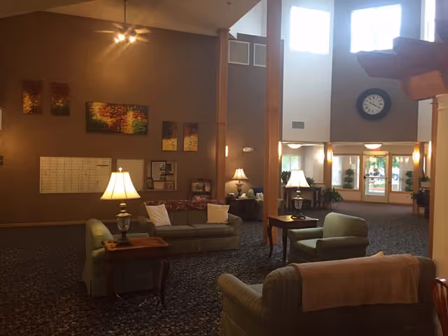 A spacious assisted living facility common area with high ceilings and large windows letting in natural light. The room features several upholstered sofas and armchairs arranged around wooden side tables with lamps. The walls are decorated with colorful abstract paintings and a large clock is mounted above the entrance doors. The carpet has a dark patterned design, and there are plants near the entrance.