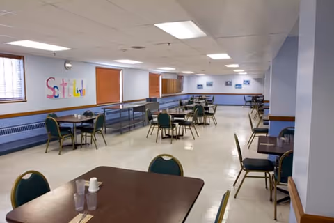 A spacious dining room with multiple tables and green cushioned chairs arranged throughout. The room has white walls with a colorful 'Scotland' sign on one wall, several windows with wooden blinds, and fluorescent ceiling lights. The floor is light-colored and there are a few framed pictures on the far wall.