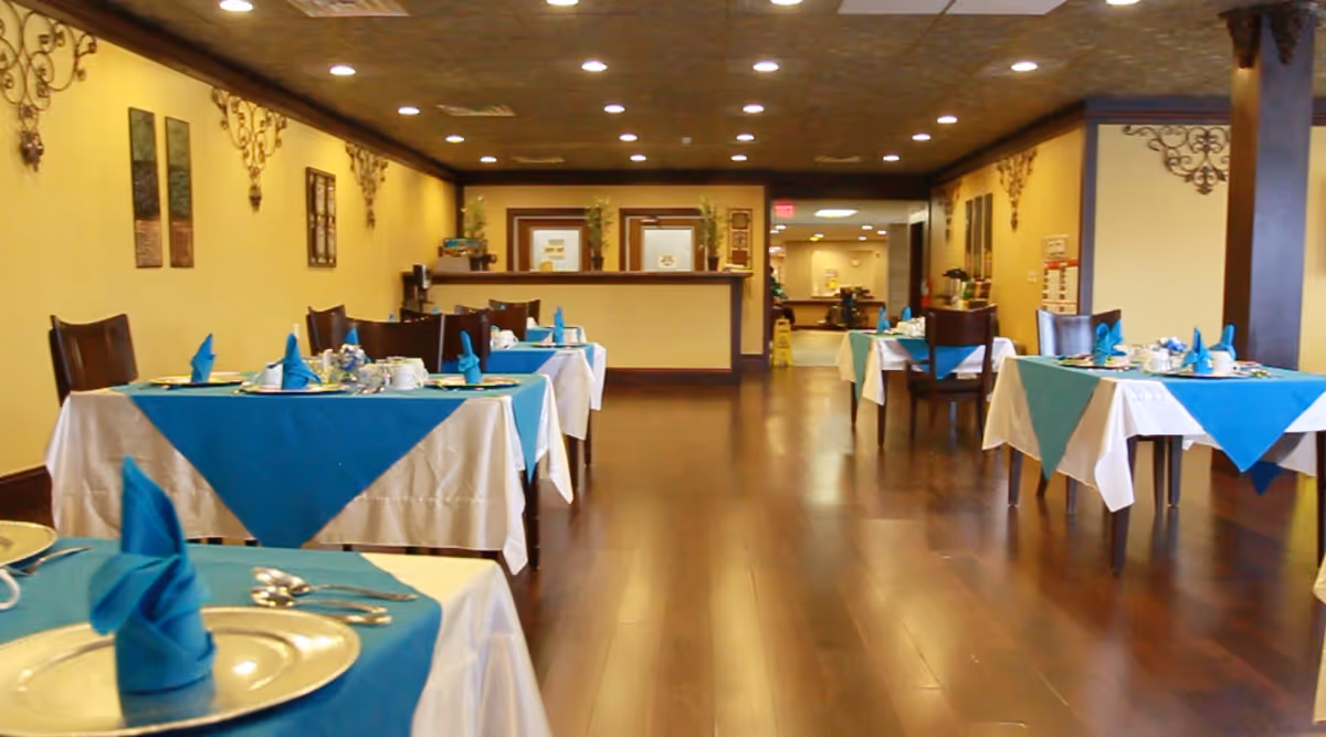 Spacious dining room with tables set with teal napkins and tablecloths, wooden floors, and a reception counter at the far end.