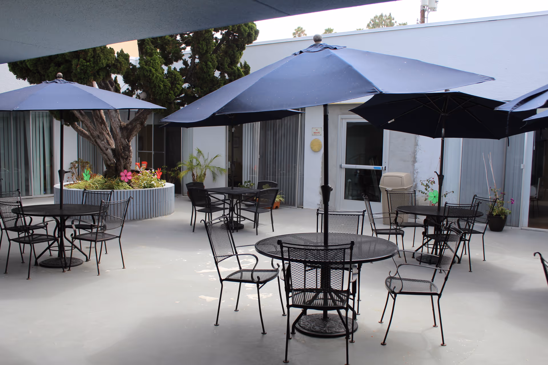 Courtyard with metal patio tables and chairs under large blue umbrellas and a central planter by sliding glass doors.