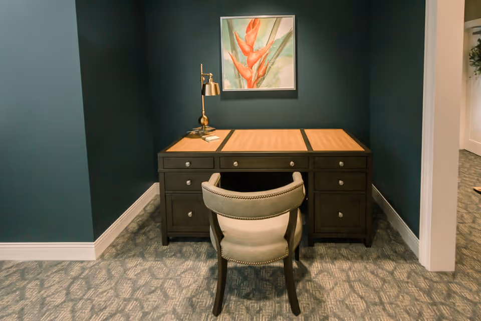 A small office nook with a dark wooden desk featuring multiple drawers and a light wood top, accompanied by a cushioned chair with nailhead trim. A brass desk lamp and a book are on the desk. The walls are painted dark teal, and a framed painting of a red flower hangs above the desk. The floor is carpeted with a patterned design.