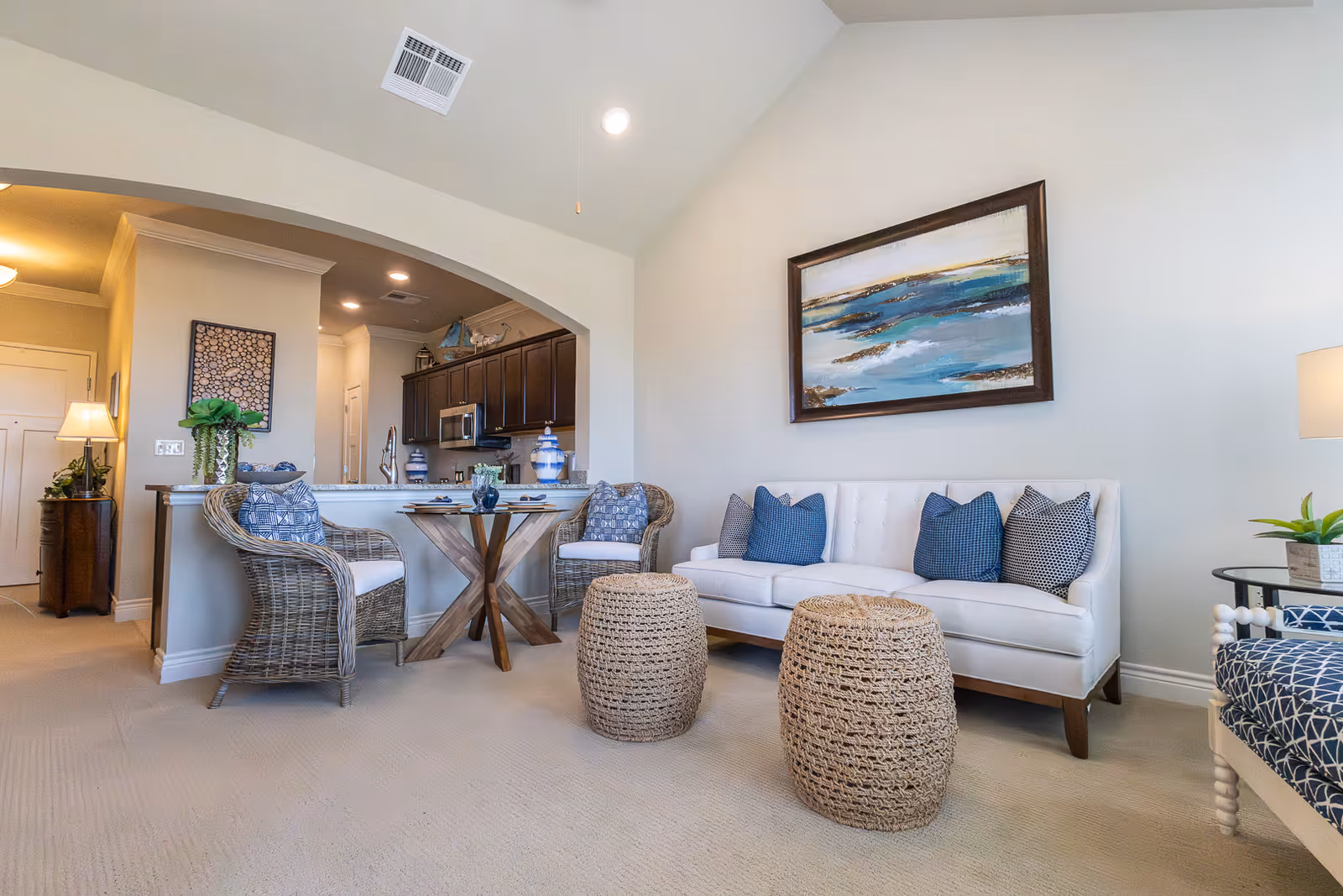 A bright and airy living room area in a senior living facility featuring a white sofa with blue and patterned pillows, two woven ottomans, a round glass-top dining table with two wicker chairs, and a kitchen with dark cabinets in the background. The walls are light-colored with a large abstract painting above the sofa, and there is a lamp and small plants adding to the cozy atmosphere.