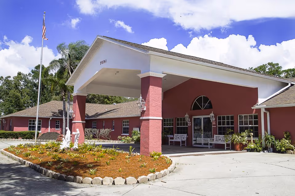 Assisted living facility exterior with covered entrance, flagpole, benches, and circular landscaped island.