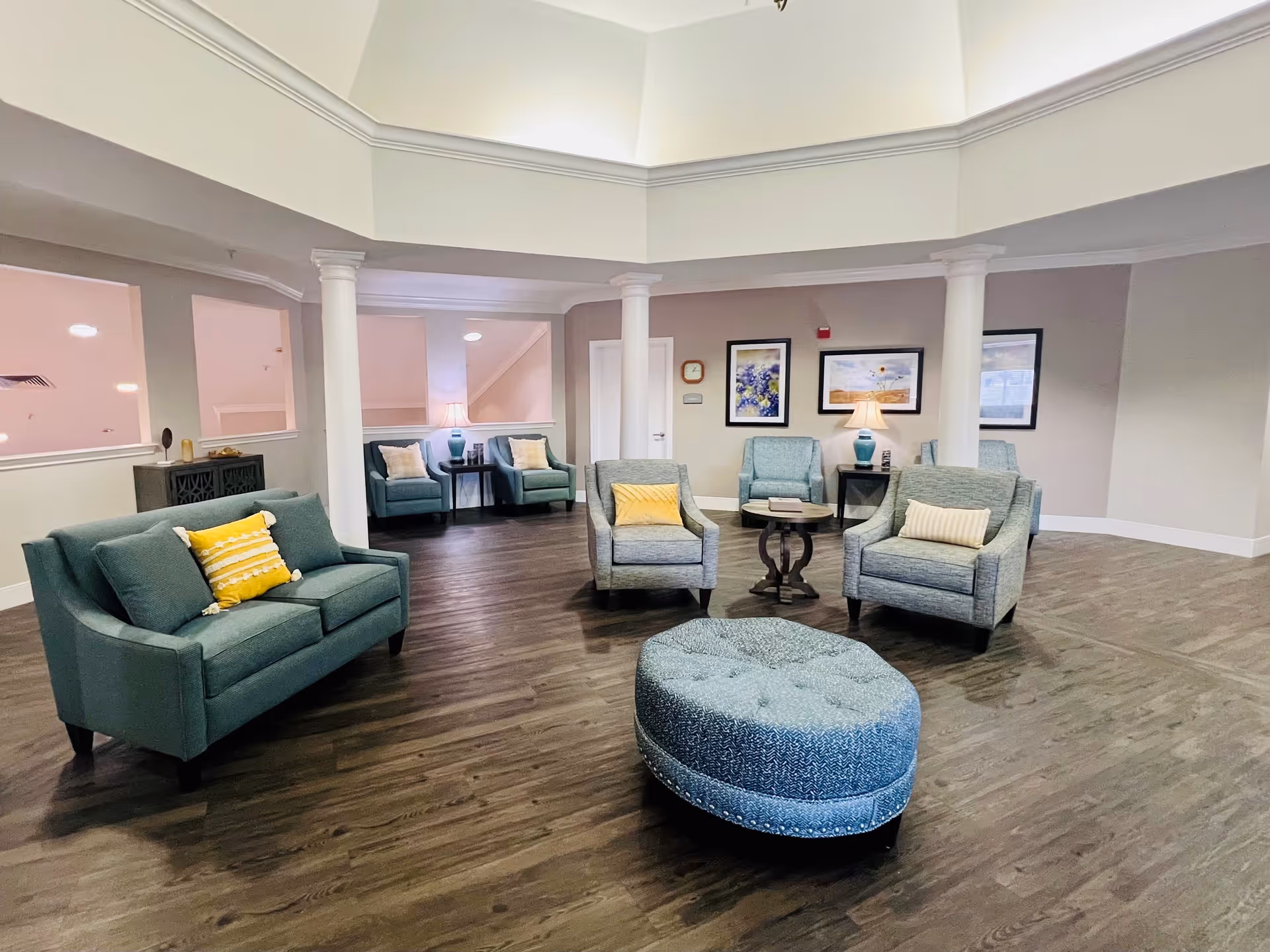 A spacious and well-lit common area with teal upholstered chairs and sofas, some with yellow and beige accent pillows. The room features dark wood flooring, white columns, and framed artwork on the walls. There is a round tufted ottoman in the center and side tables with lamps, creating a cozy and inviting atmosphere.