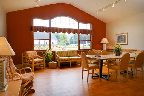 A bright and spacious common area with wooden flooring, featuring a table with six chairs, two armchairs, a wooden bench, and a rocking chair. Large windows with a red valance allow natural light to fill the room. There are plants and framed artwork on the walls, creating a welcoming atmosphere.