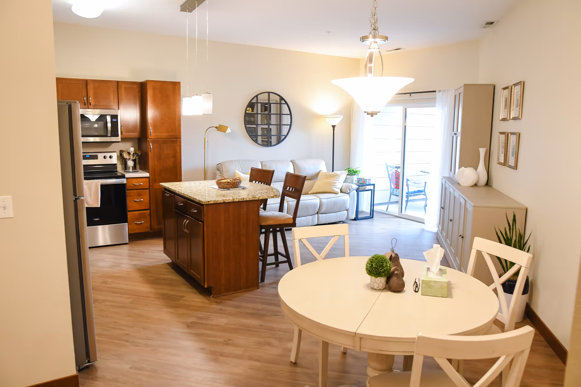 Open-plan living area with a round dining table in the foreground, a kitchen island and stainless-steel appliances to the left, and a sofa near a sliding glass door in the background.
