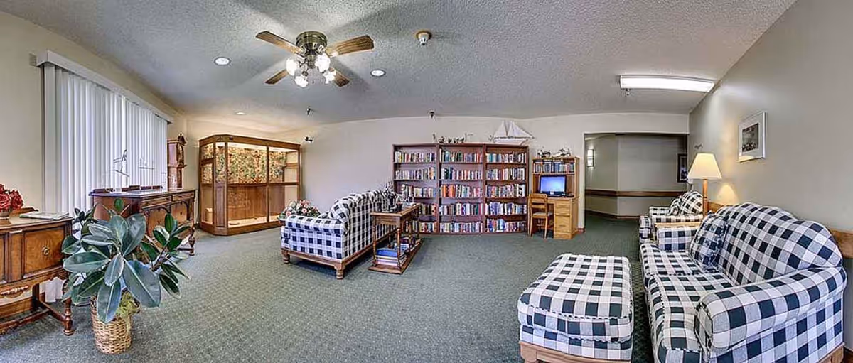 A senior living community common area with checkered upholstered sofas and ottoman, a wooden bookshelf filled with books, a small desk with a computer, a ceiling fan with lights, and a potted plant near a window with vertical blinds.