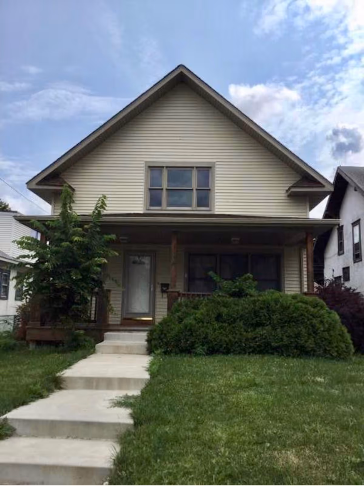 Two-story beige house with a covered front porch, concrete walkway, and lawn with shrubs.