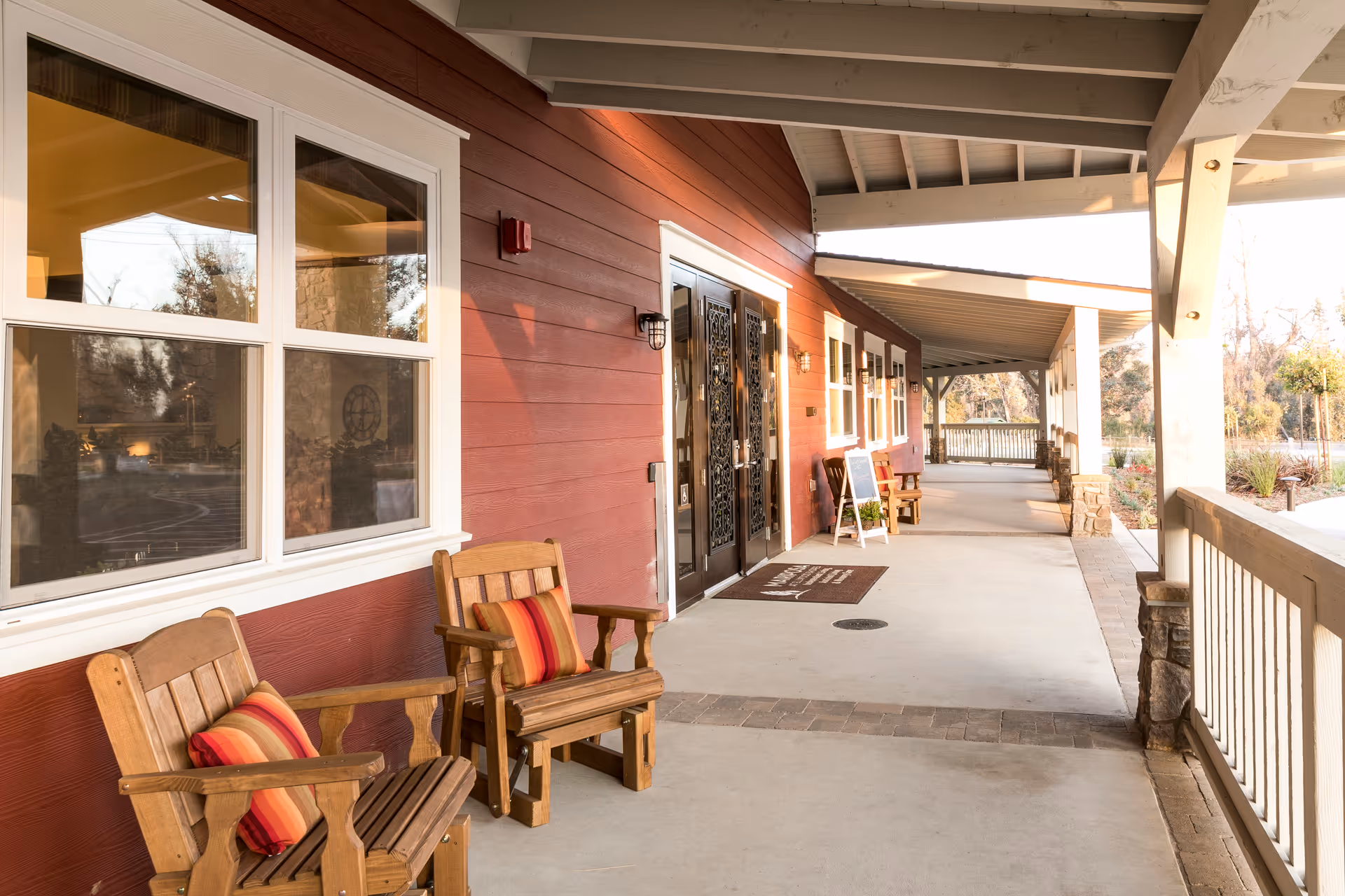 Covered outdoor porch area with wooden chairs featuring red and orange striped cushions, red siding wall with windows, decorative double doors, and a railing overlooking a garden area.