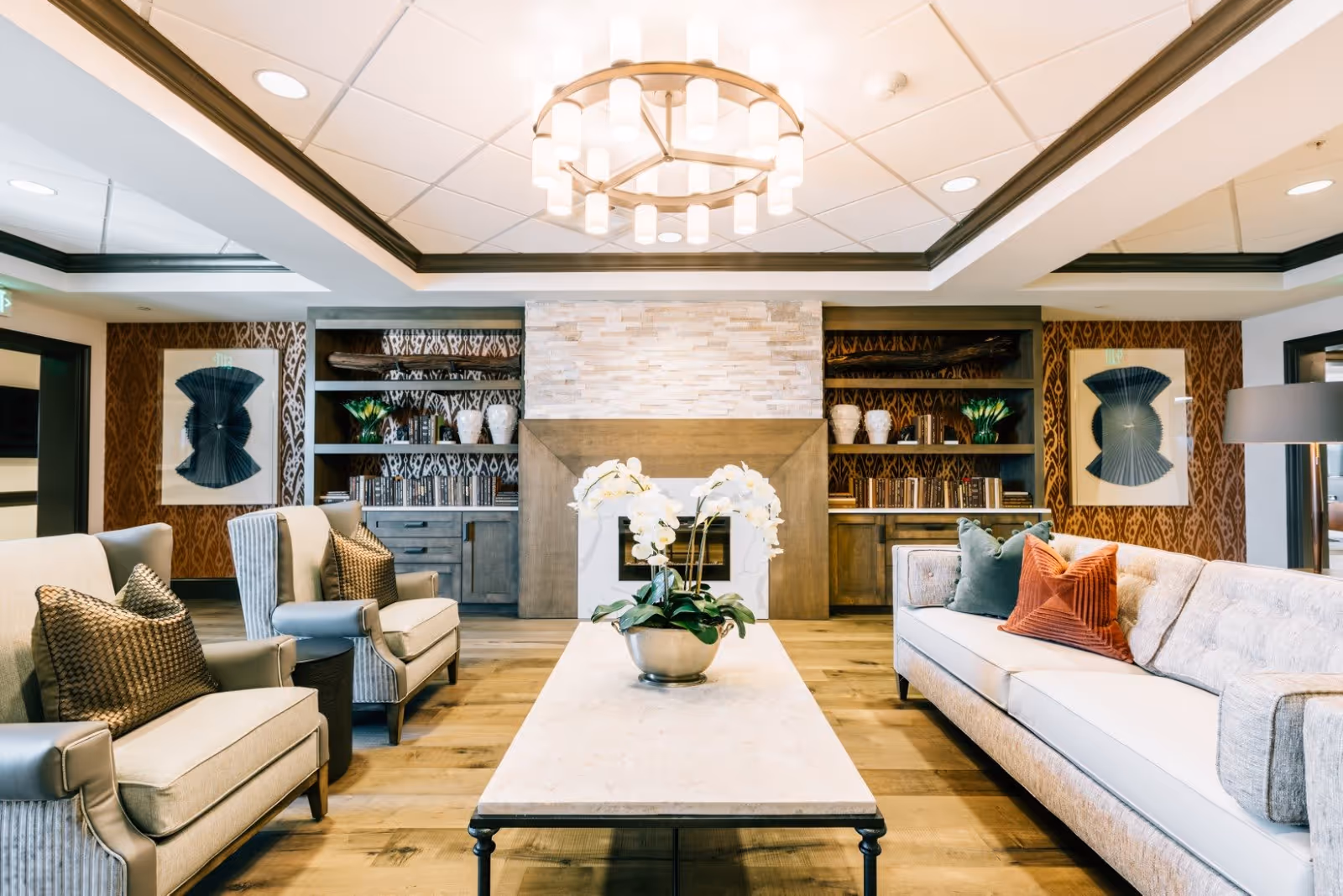 A bright and elegant living room with a large beige sofa adorned with colorful pillows, two armchairs with patterned cushions, a rectangular coffee table with a potted white orchid centerpiece, built-in wooden shelves filled with books and decorative items, and a modern chandelier hanging from the ceiling.