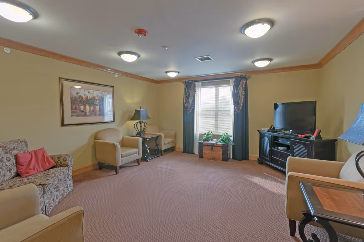 Well-lit communal living room with upholstered chairs and sofa, a TV on a console, lamps, and a curtained window.