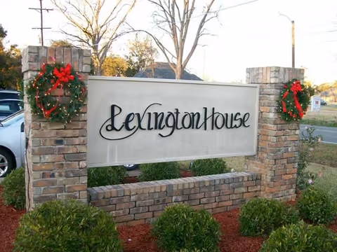 A brick signpost with a rectangular white sign displaying the text 'Lexington House' in black cursive font. The signpost is decorated with two Christmas wreaths with red bows, one on each brick pillar. There are green bushes and red mulch around the base of the sign, and a street with trees and cars is visible in the background.