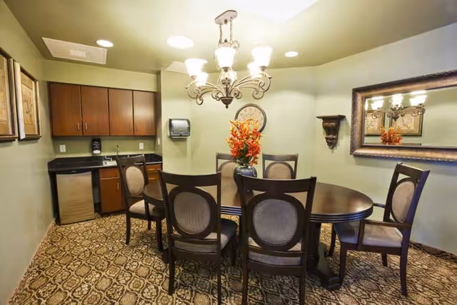 A dining room with a round wooden table surrounded by six upholstered chairs. A chandelier with multiple lights hangs above the table. On the table is a vase with orange flowers. The room has light green walls, a large mirror on one wall, a clock, and a small kitchenette area with wooden cabinets, a sink, and a mini fridge. The floor is covered with patterned carpet.