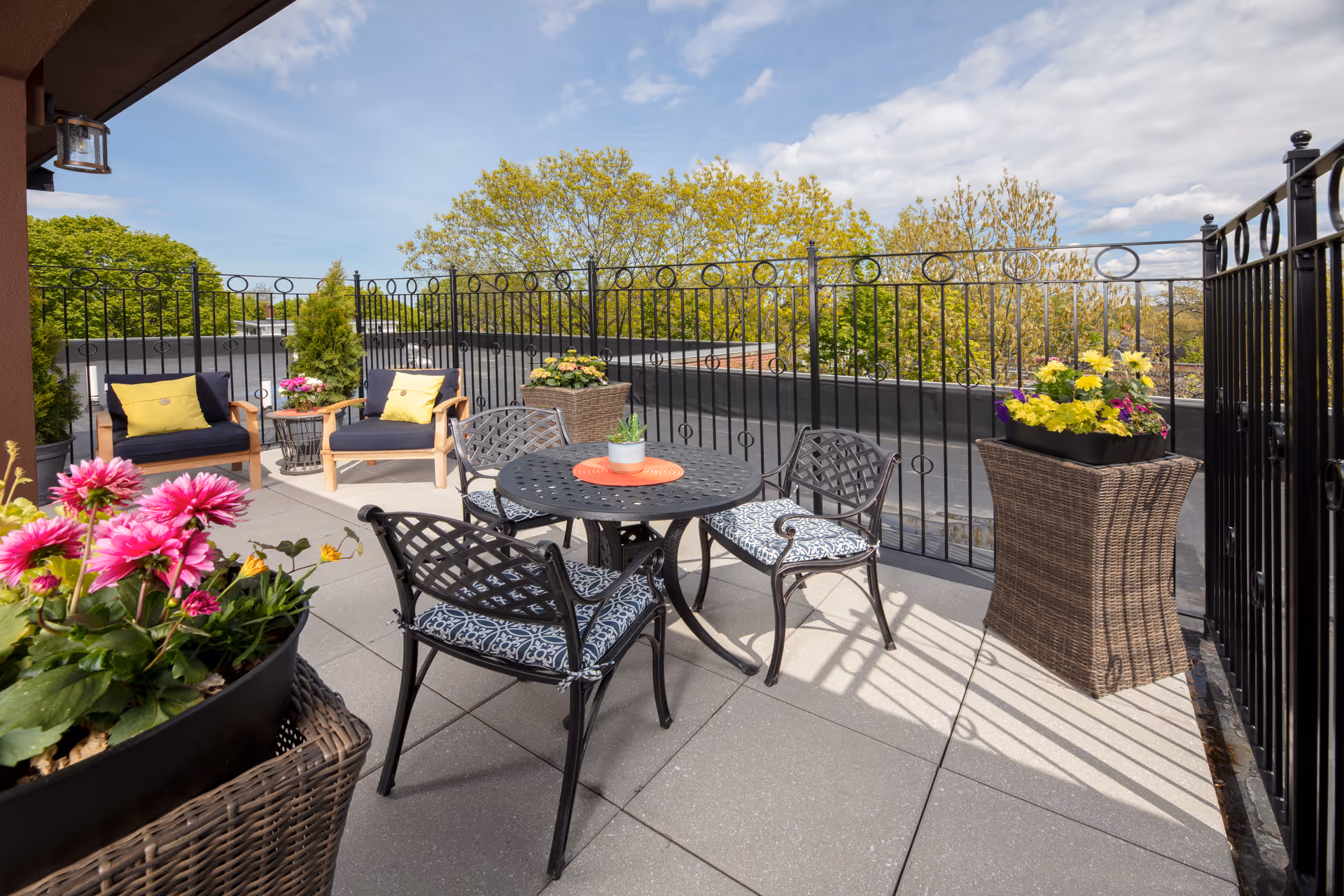Rooftop patio with a round metal table and chairs, cushioned armchairs, and potted flowers along a decorative metal railing under a blue sky.