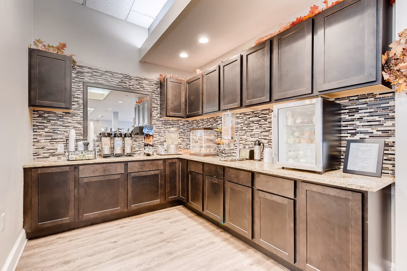 A kitchenette area with dark wood cabinets, a granite countertop, and a mosaic tile backsplash. The countertop holds coffee dispensers, cups, a small refrigerator with snacks, and a framed menu. The space is well-lit with recessed ceiling lights and decorated with autumn leaves on top of the cabinets.