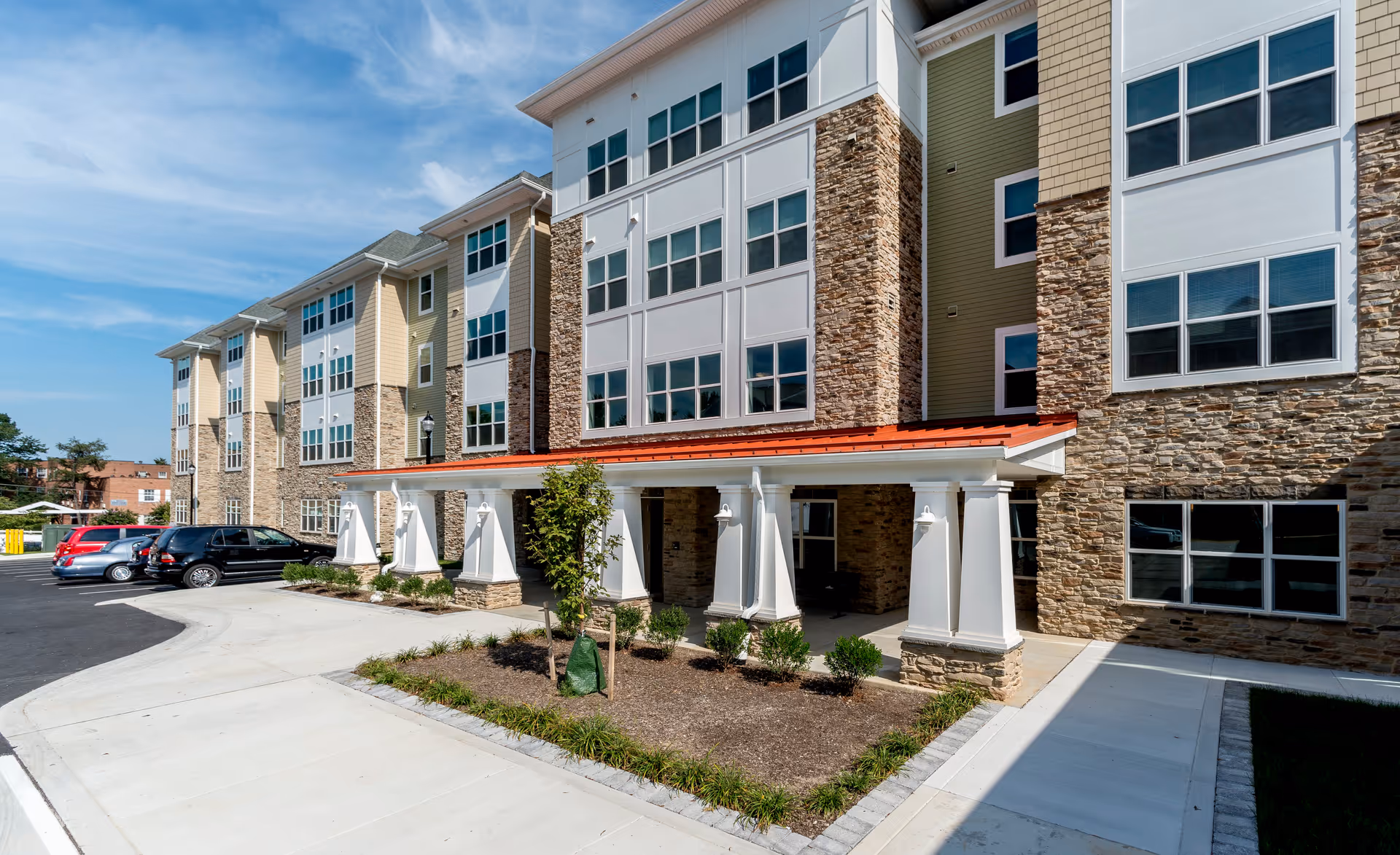 Exterior view of a multi-story senior living facility building with stone and siding facade, large windows, a covered entrance with white columns, a landscaped area with small plants and a tree, and a parking lot with several cars under a blue sky.