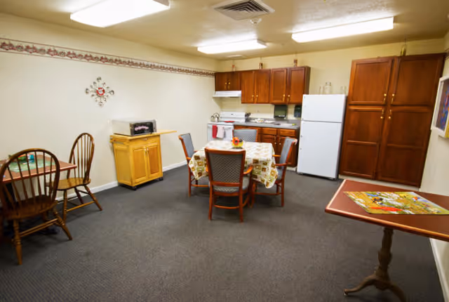 A communal dining room with a round table and chairs, a small kitchenette with cabinets, refrigerator and stove, and additional tables around the room.