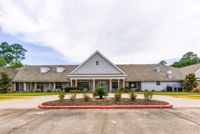Front exterior view of a single-story senior living facility building with a gabled roof, a covered entrance supported by columns, landscaped bushes and plants in front, and a circular driveway with a red curb.