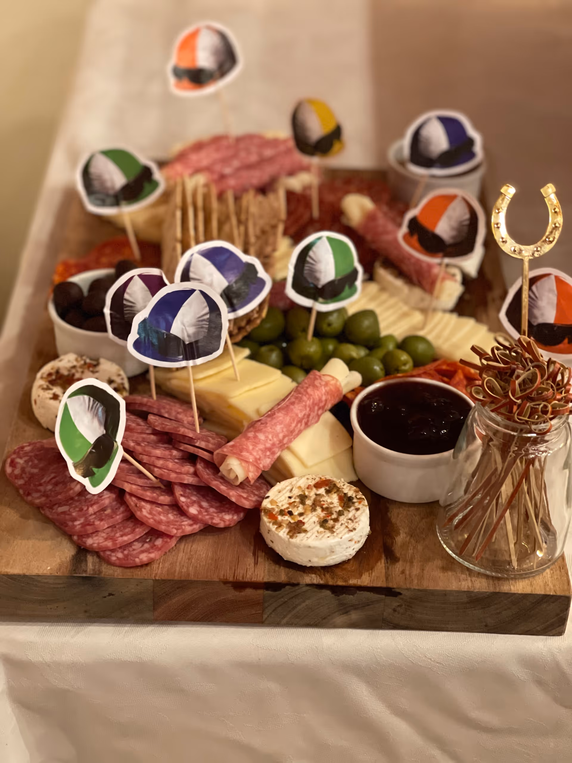 A wooden charcuterie board featuring an assortment of sliced salami, rolled meats, cheeses, green olives, crackers, and small bowls of dark jam and olives. The board is decorated with small paper cutouts of jockey helmets on toothpicks and a gold horseshoe-shaped pick holder with cocktail picks.