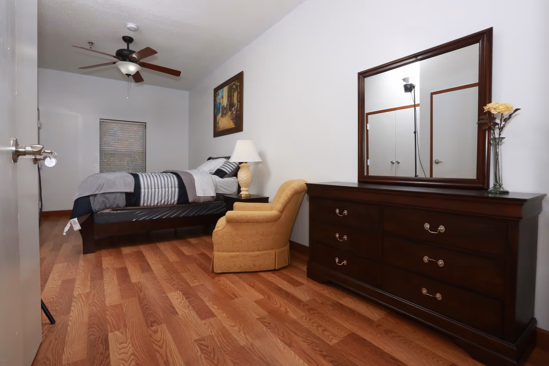 A furnished bedroom with a bed, yellow armchair, wooden dresser with mirror, and a ceiling fan over hardwood floors.
