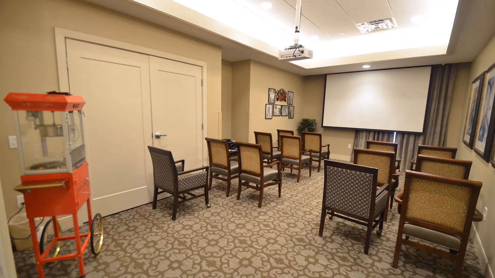 A small theater room with rows of chairs facing a large projector screen. There is a red popcorn machine on the left side near double doors, patterned carpet, framed pictures on the walls, and a ceiling-mounted projector.