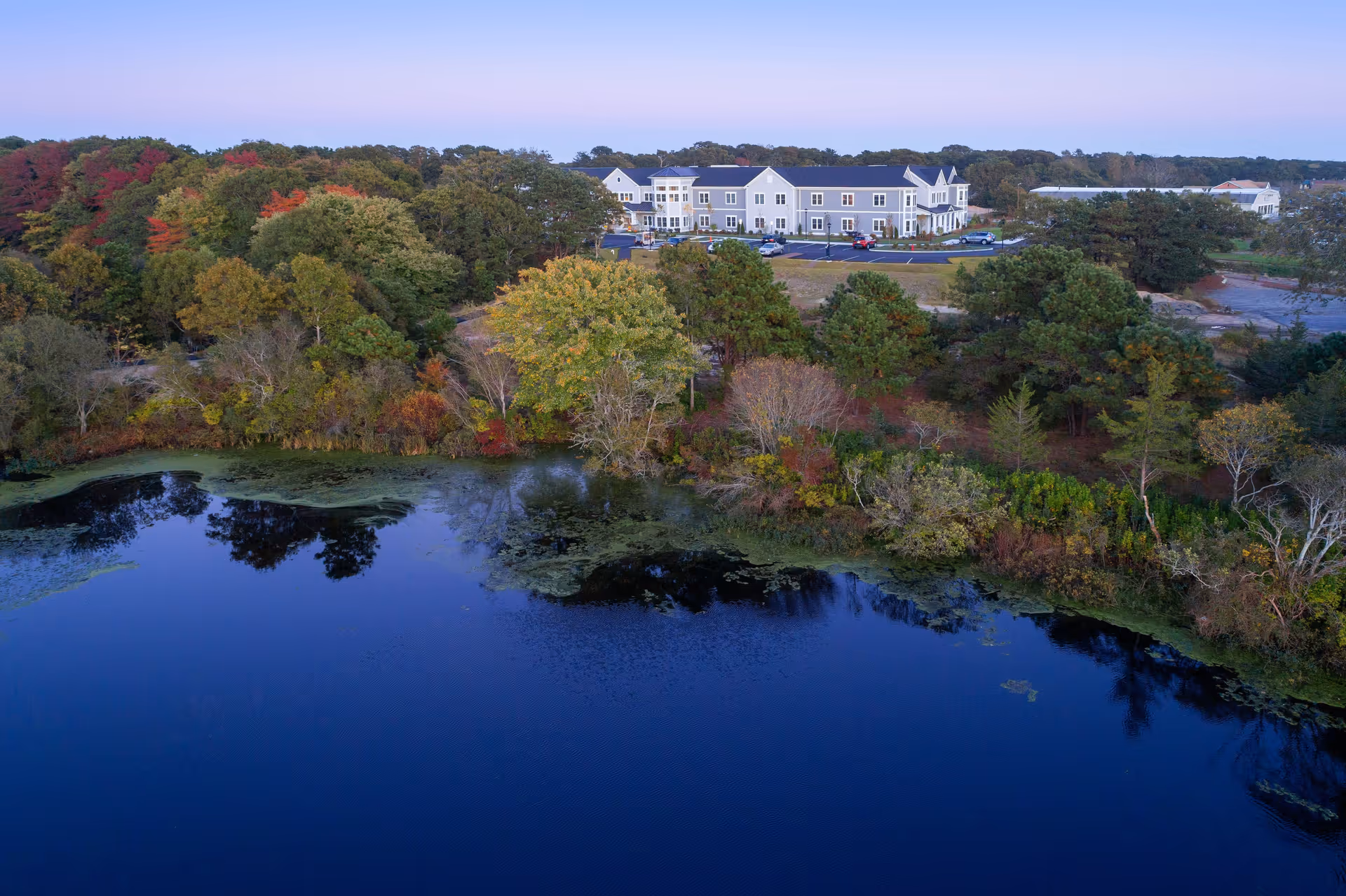 Aerial view of a senior living facility named Maplewood at Mill Hill surrounded by trees with autumn foliage, adjacent to a calm body of water reflecting the trees and sky.