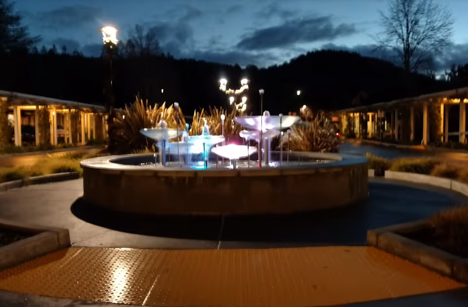 A circular illuminated fountain in a courtyard at dusk with surrounding walkways and lampposts.