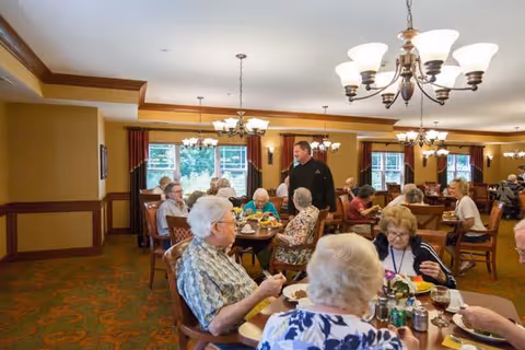 A dining room at Shoreview Senior Living with several elderly residents seated at tables enjoying a meal. A staff member is standing and interacting with the residents. The room is warmly lit with chandeliers, has large windows with red curtains, and wooden trim along the walls.