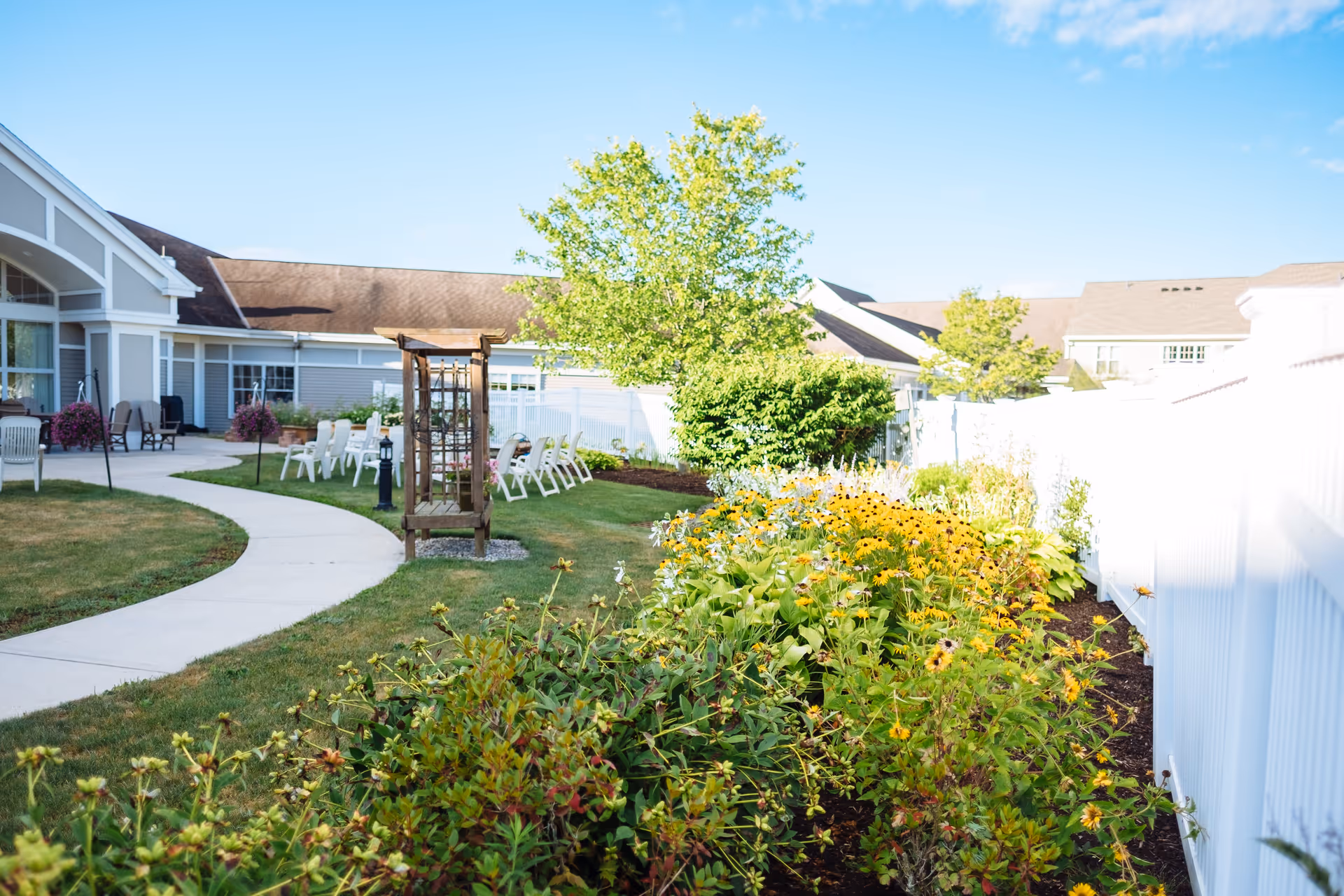 Outdoor garden area at The Terrace at The Glen with a curved concrete pathway, green lawn, flower beds with yellow flowers, a wooden trellis, white fence, and patio chairs near the building under a clear blue sky.
