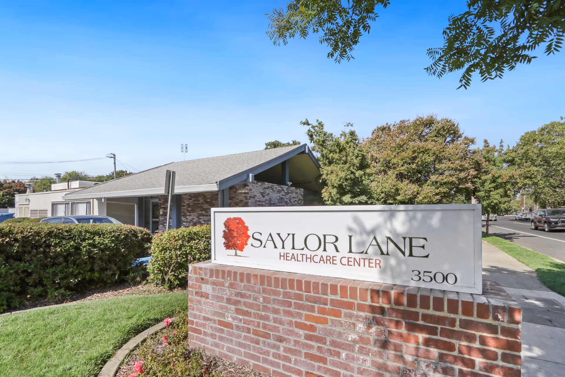 Exterior view of Saylor Lane Healthcare Center showing a brick sign with the facility's name and address 3500, surrounded by greenery and trees under a clear blue sky.