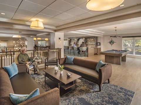 A spacious senior living facility common area with two brown sofas and a coffee table in the foreground. An elderly man is sitting on one sofa reading a newspaper. In the background, there is a bar area with stools and a pool table near large windows letting in natural light.