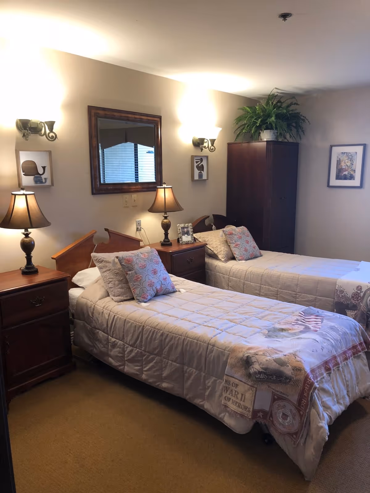 A cozy bedroom with two single beds, each with a beige quilt and decorative pillows. Between the beds is a wooden nightstand with a lamp and a framed photo. Above the nightstand is a large mirror on the wall, flanked by two wall-mounted light fixtures. A dark wooden wardrobe with a potted plant on top is in the corner, and a framed picture hangs on the adjacent wall. The room has warm lighting and a carpeted floor.
