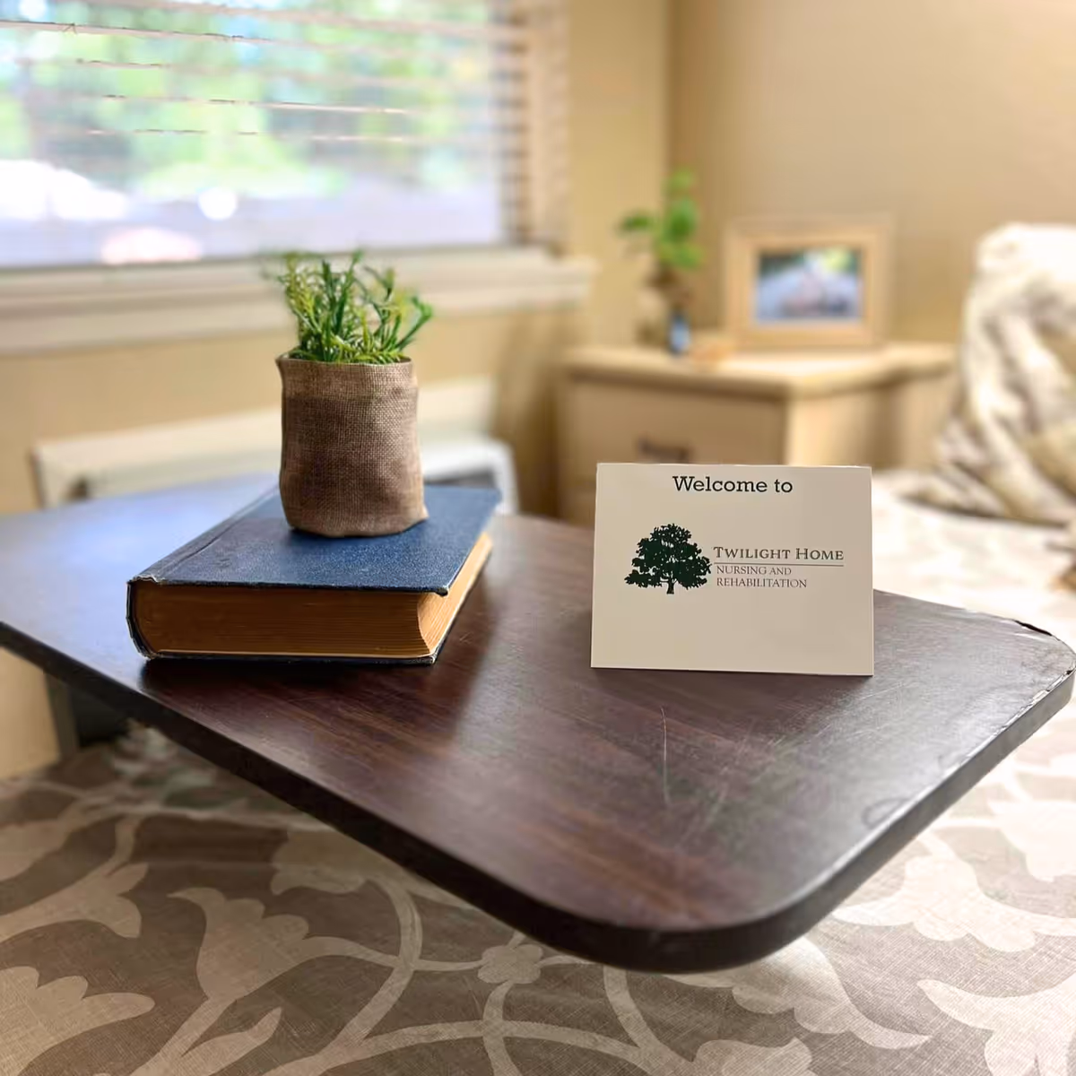A tray table in a bright bedroom displays a closed book, a small potted plant, and a 'Welcome to Twilight Home Nursing and Rehabilitation' card.