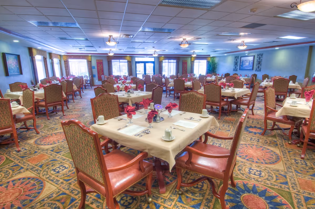 A spacious dining room with multiple tables covered in beige tablecloths, each set with cups, utensils, and red folded napkins. The room features patterned carpet flooring, upholstered chairs, and large windows letting in natural light.