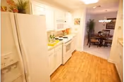 Bright kitchen with white cabinets, white refrigerator, stove, microwave, and wooden flooring. The kitchen opens into a dining area with a round table and chairs, and a plant is visible in the corner.