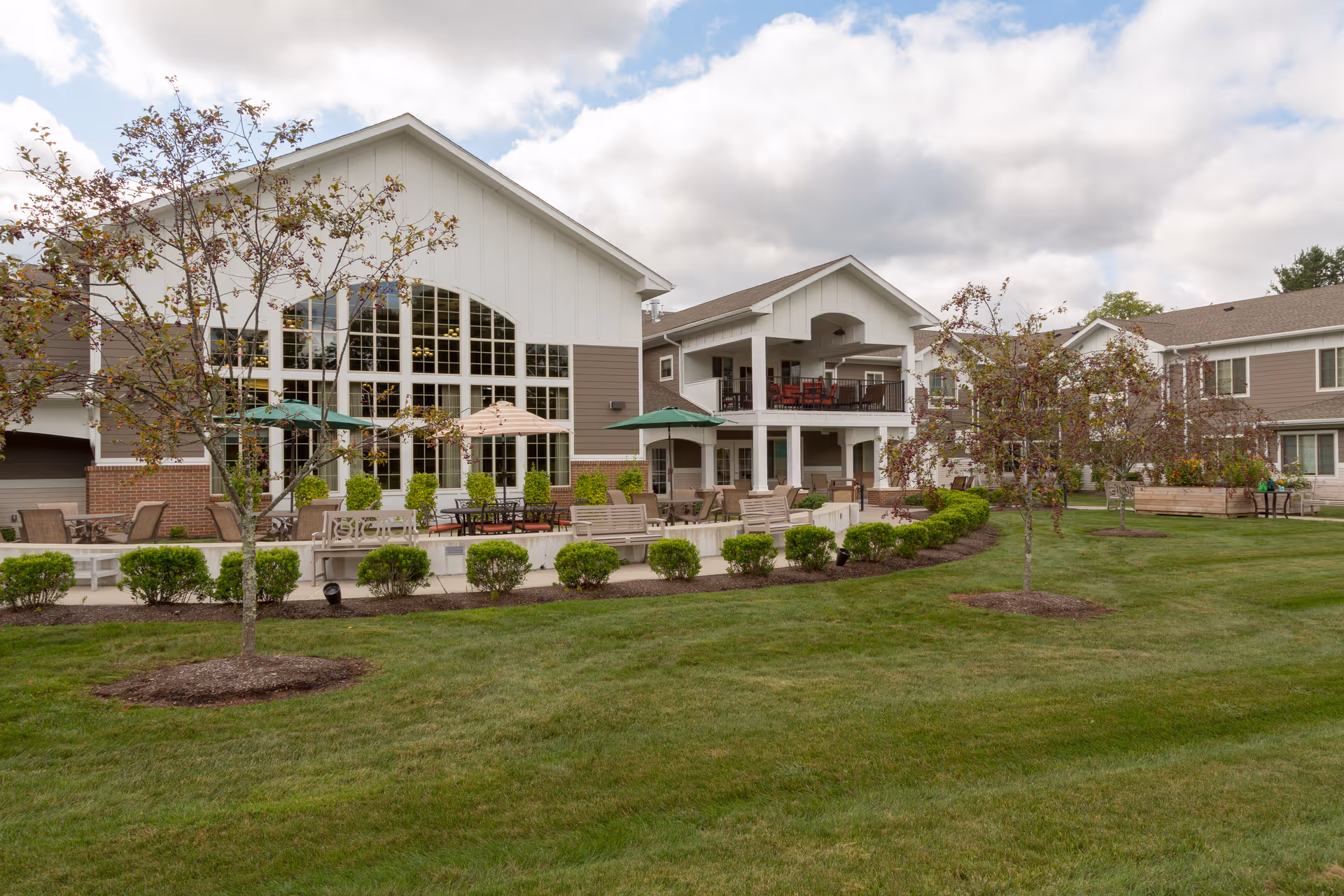 Exterior view of a two-story senior living building with large windows, outdoor patio seating and umbrellas overlooking a landscaped lawn.