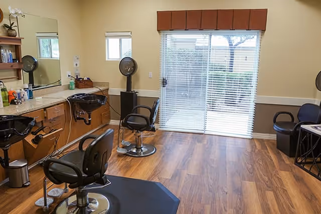 Interior view of a hair salon area in a senior living facility with two black salon chairs in front of two black hair washing sinks. There is a large mirror on the wall, shelves with hair products, a fan, and a sliding glass door with blinds leading outside. The floor is wood and the walls are painted beige with a white chair rail.