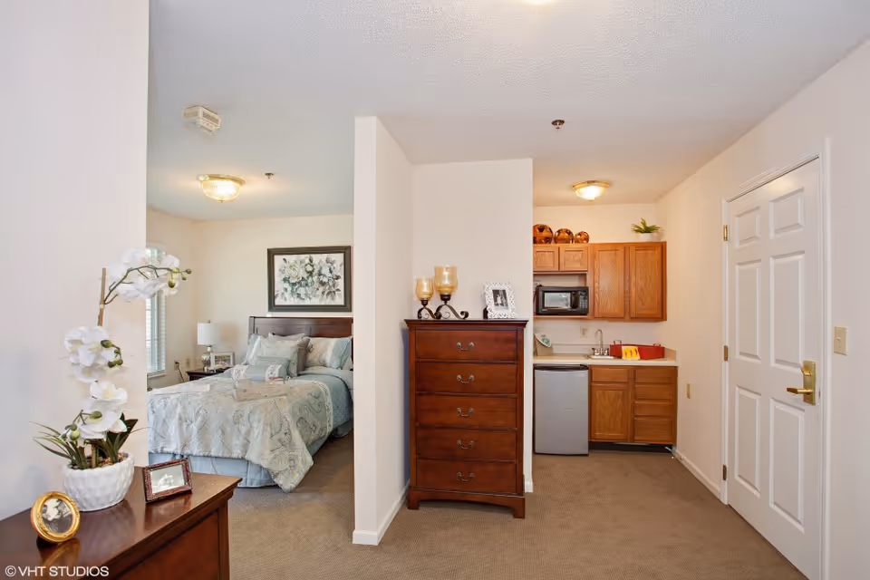 View of a senior living facility apartment showing a bedroom with a bed, nightstand, and lamp on the left, a wooden chest of drawers in the center, and a small kitchenette with cabinets, microwave, mini fridge, and sink on the right. The walls are light-colored and the floor is carpeted.