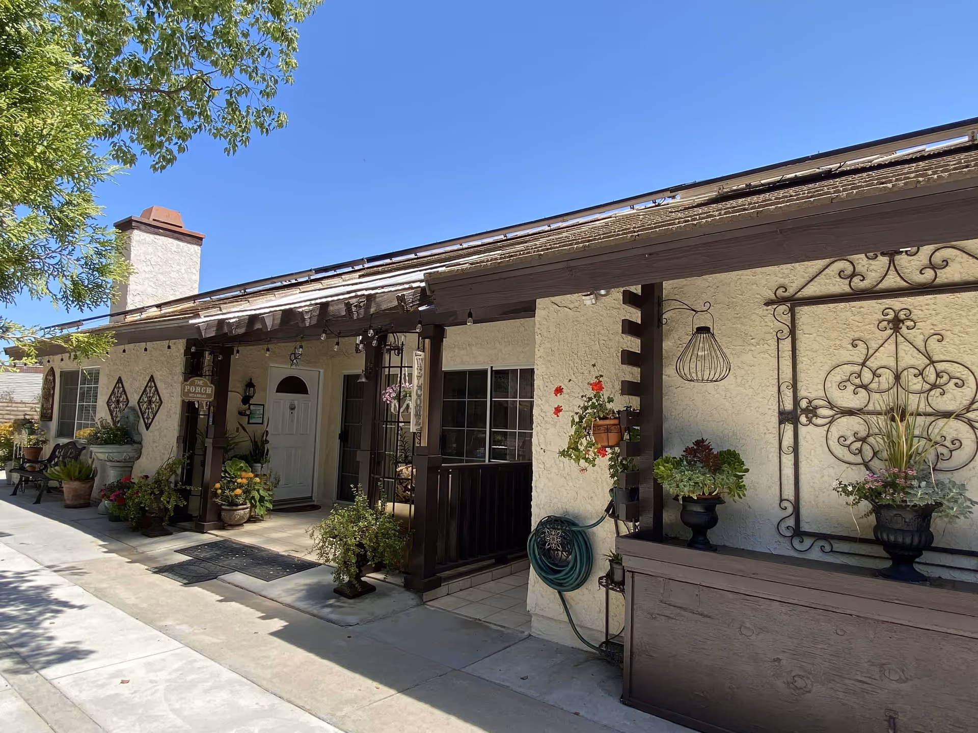 Exterior view of a single-story building with a covered porch area featuring wooden beams and various potted plants. The building has a textured cream-colored facade, a white door, and decorative metal wall art. A garden hose is coiled on the wall, and there is a clear blue sky above.