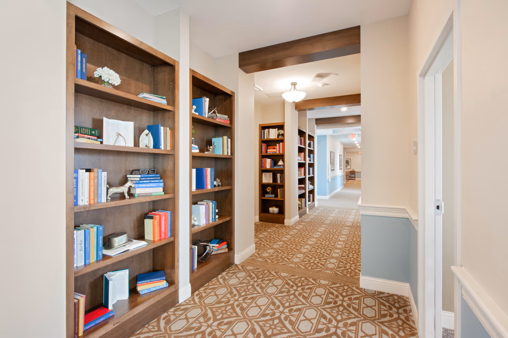 A well-lit hallway in a senior living facility with built-in wooden bookshelves filled with books and decorative items. The hallway has patterned carpet flooring, light-colored walls with a blue lower half, and ceiling light fixtures. The corridor extends into the distance with more bookshelves and framed pictures on the walls.