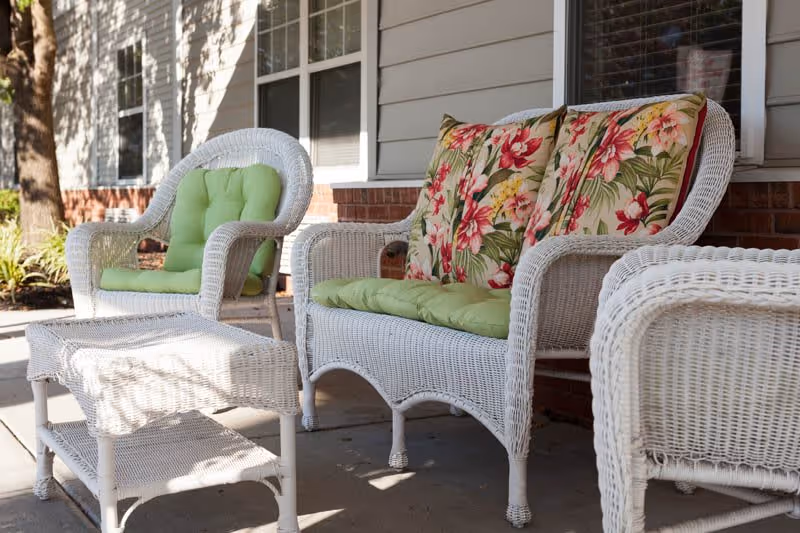 Outdoor seating area with white wicker furniture including a loveseat with floral and green cushions, a chair with a green cushion, and a matching wicker table, set on a concrete patio in front of a building with beige siding and windows.