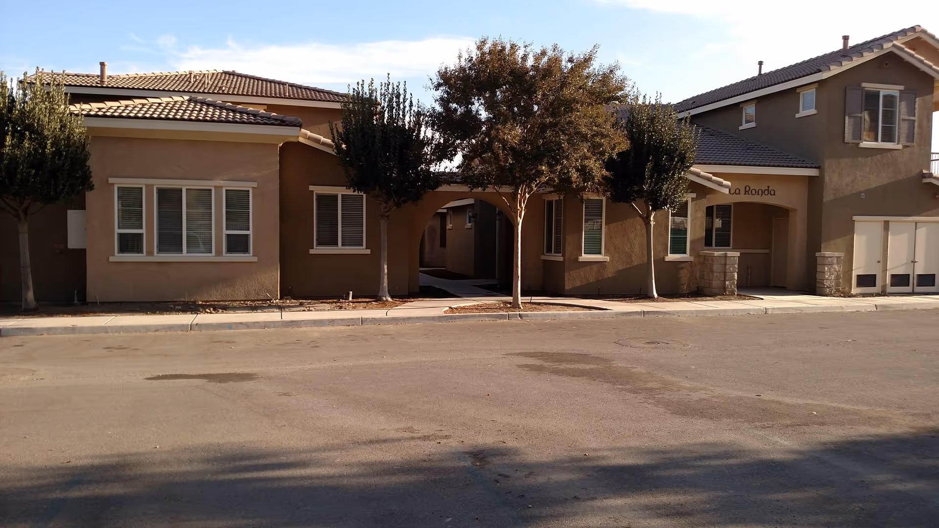 Exterior view of a residential building with beige walls and tiled roofs, featuring several windows and an archway in the center. There are four trees planted along the sidewalk in front of the building, and the street is visible in the foreground. The building has a sign that reads 'La Ronda'.