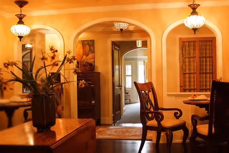 A warmly lit interior room with classic wooden furniture including a table and chairs. There are arched doorways and windows with curtains, a vase with flowers on a wooden surface, and a glimpse into another room with a window and seating area.