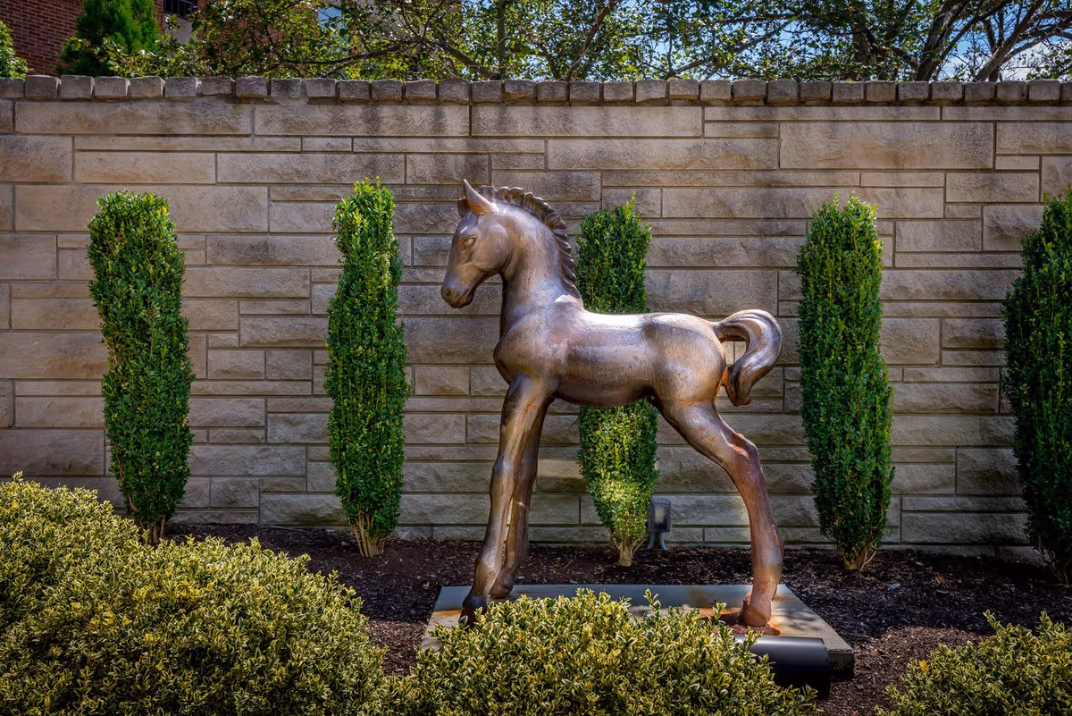 A bronze statue of a young horse standing on a concrete base in a garden area with neatly trimmed bushes and tall green shrubs against a stone wall background.