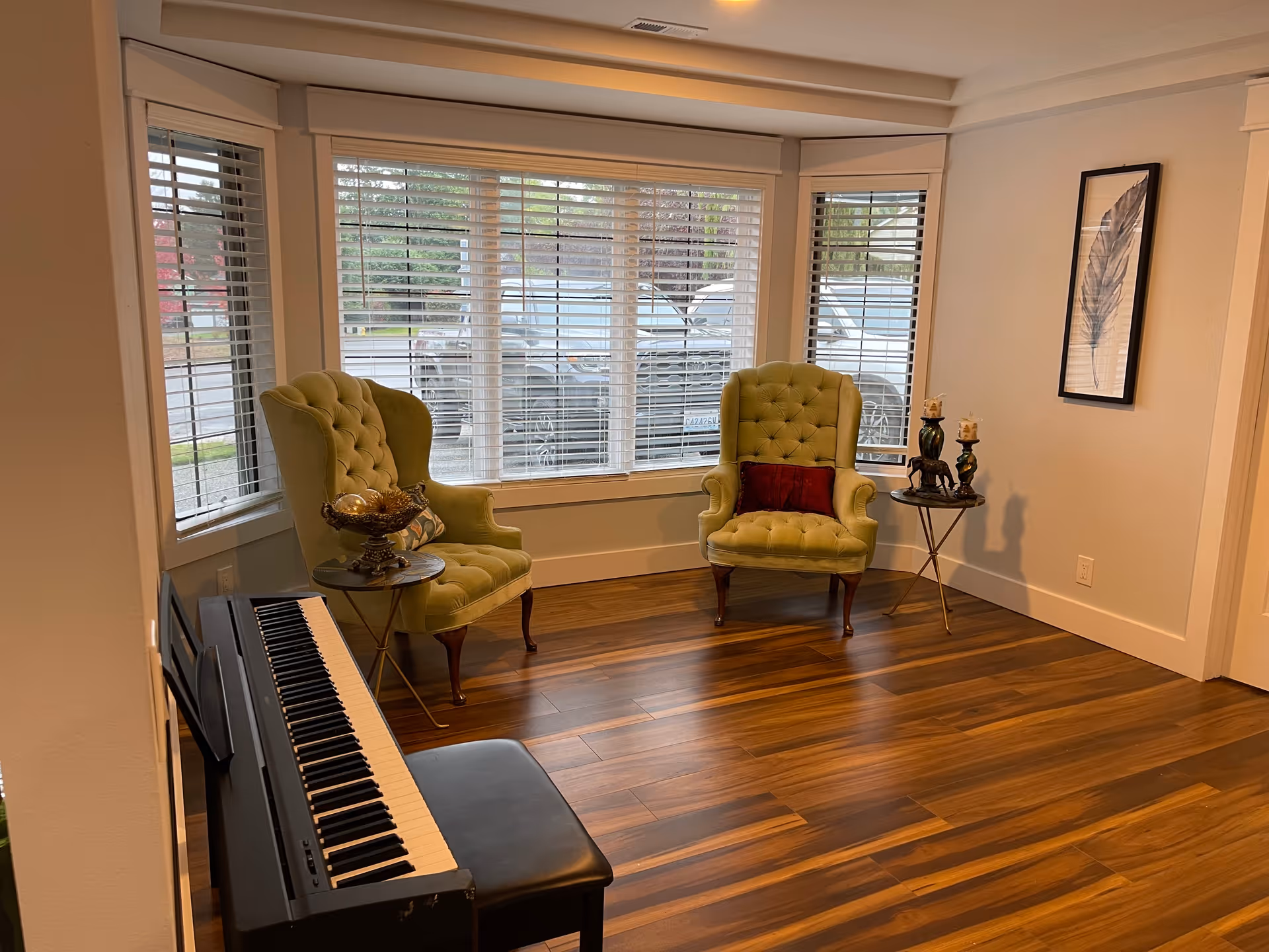 Sunlit sitting area with two green tufted armchairs, a keyboard piano, and a bay window with blinds.